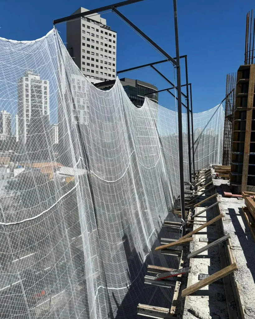 Fachada de edifício em construção com Sistema de Limitador de Quedas de Altura (SLQA) da R2K instalado, mostrando as redes de proteção periférica e suportes metálicos integrados à laje.