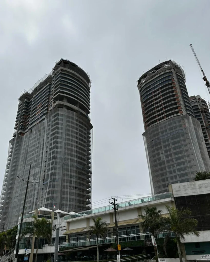 Vista ampla de um edifício em construção com múltiplos pavimentos protegidos por redes de segurança e sistemas de proteção periférica R2K, evidenciando a organização e segurança do canteiro.