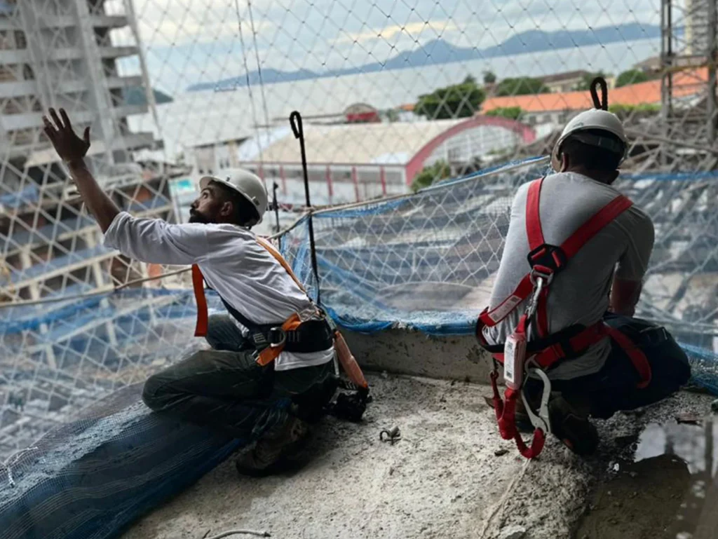 Equipe técnica da R2K realizando a instalação de rede de proteção piso a piso em estrutura de edifício, utilizando cintos de segurança e seguindo as normas NR-18 e NR-35.