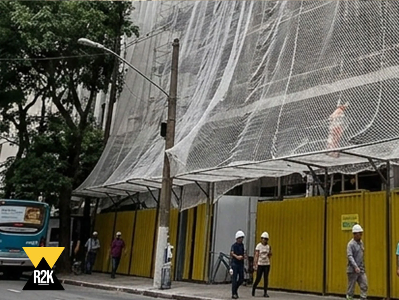 Uma visão geral de um grande canteiro de obras cercado por tapume amarelo e coberto por uma rede de segurança branca. Pedestres e trabalhadores andam na calçada. Uma placa de aviso em português está visível.